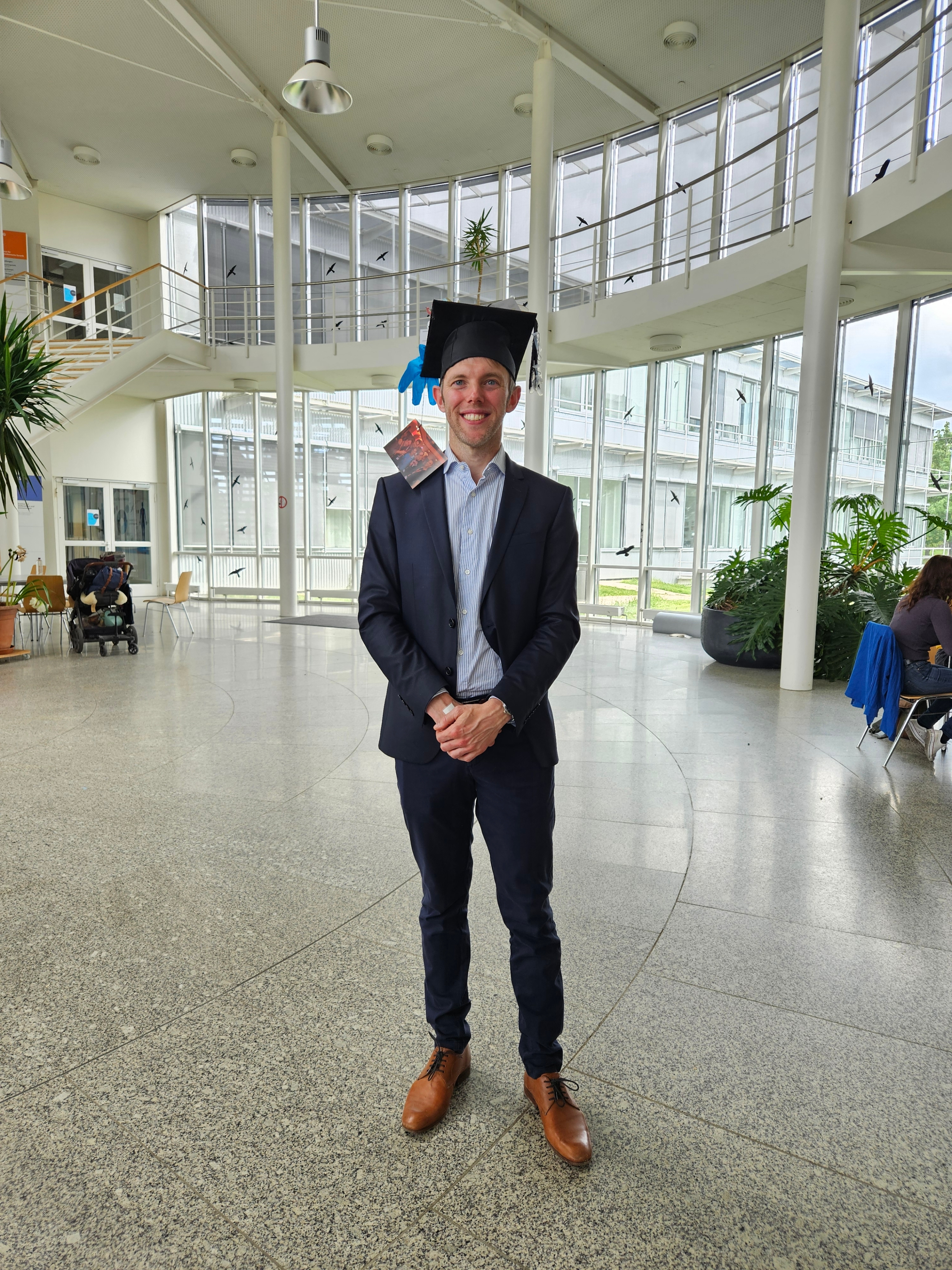 A man in a suit is standing alone, smiling. He is wearing a typical "PhD passed" hat